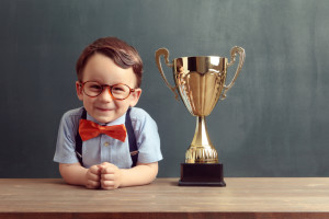 A cute, smiling 2-3 years old boy is standing behind a wooden table with a golden trophy on it. Little boy is wearing an orange bow tie, red glasses and blue trousers with suspenders.